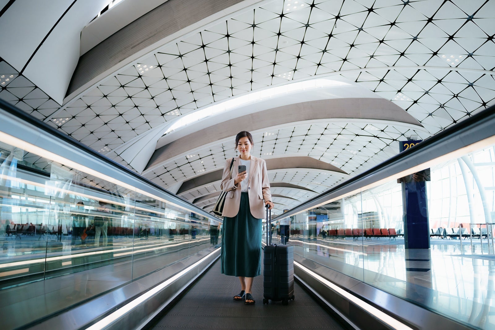Young Asian businesswoman with suitcase using smartphone while walking on moving walkway at airport terminal. Business traveller. Lifestyle and technology. Travel and vacation concept